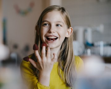 Ein Mädchen mit langen Haaren trägt ein gelbes Oberteil und lächelt, während es Erdbeeren auf ihren Fingern hält. Im Hintergrund sind verschwommene Küchenutensilien zu sehen.