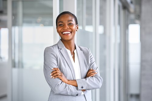 Eine lächelnde Frau in einem grauen Anzug steht mit verschränkten Armen in einem modernen Büro.