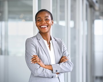Eine lächelnde Frau in einem grauen Anzug steht mit verschränkten Armen in einem modernen Büro.