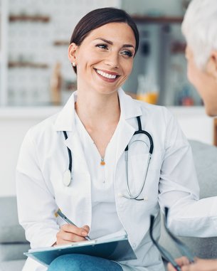 Eine Ärztin in einem weißen Kittel sitzt auf einem Sofa und spricht mit einer älteren Patientin. Die Ärztin hält einen Notizblock und lächelt, während die Patientin aufmerksam zuhört.