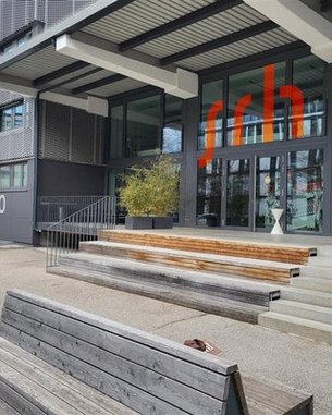 Entrance to a modern building with large windows, wooden steps, and a wooden bench in the foreground. The number 10 is clearly visible, and an orange logo is mounted on the wall.