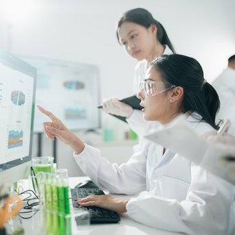A scientist points to data on a computer screen while a colleague takes notes. In the background, test tubes are visible.
