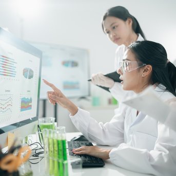 Eine Wissenschaftlerin zeigt auf Daten auf einem Computerbildschirm, während eine Kollegin Notizen macht. Im Hintergrund sind Reagenzgläser sichtbar.