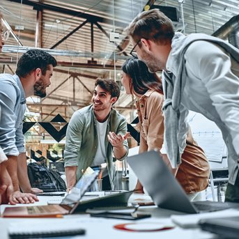 Sechs Personen in einem modernen Büro diskutieren lebhaft um einen Tisch mit Laptops und Notizen.