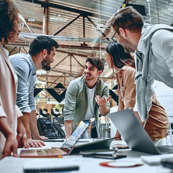 Sechs Personen in einem modernen Büro diskutieren lebhaft um einen Tisch mit Laptops und Notizen.