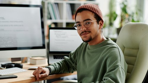 Junger Mann mit Brille und Mütze sitzt an einem Schreibtisch, umgeben von Computern und Notizen, und schaut in die Kamera.
