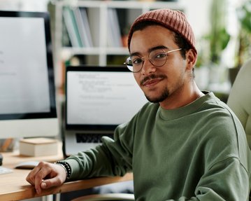 A young man with glasses and a hat sits at a desk, surrounded by computers and notes, looking into the camera.