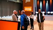 Menschen stehen in einem Konferenzraum, während eine Präsentation vorbereitet wird. Im Hintergrund ist ein Bildschirm mit dem Text "Internationale Konferenz zu nachhaltiger Stadtentwicklung" sichtbar. Stühle sind für die Teilnehmer aufgestellt.