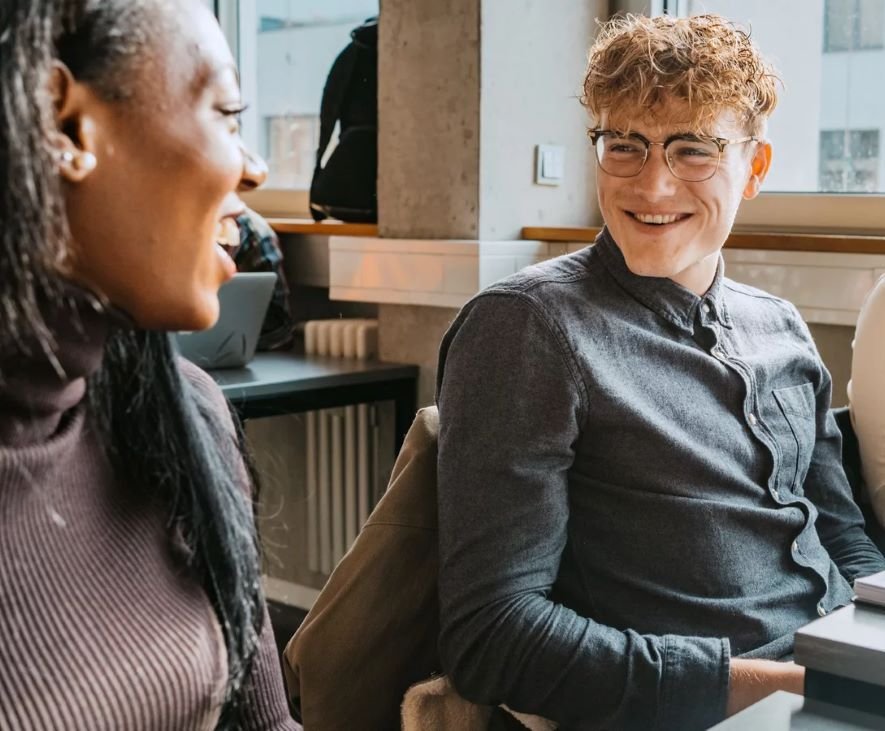 Drei junge Menschen sitzen an einem Tisch, lächeln und unterhalten sich. Bücher und Notizen liegen vor ihnen, während sie in einer hellen, modernen Umgebung lernen.