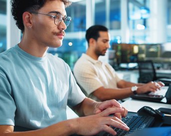 A young man with glasses is typing intently at a computer, while another man in the background is working at his own computer.