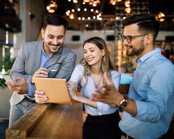 Drei Personen in einem modernen Café schauen auf ein Tablet und diskutieren. Sie wirken engagiert und freundlich.
