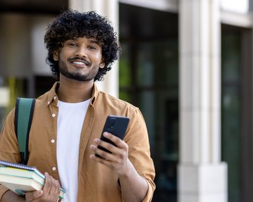 A young man with curly hair is holding a smartphone in one hand and books under his arm while smiling. He is wearing a backpack.
