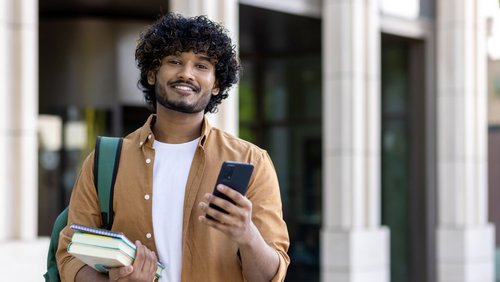Ein junger Mann mit lockigem Haar hält ein Smartphone in der Hand und Bücher unter dem Arm, während er lächelt. Er trägt einen Rucksack.