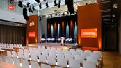 Konferenzraum mit einer Bühne, ausgestattet mit einem Rednerpult und mehreren Stühlen. Im Hintergrund sind orangefarbene Wände mit Beleuchtung und Bildschirmen, die Informationen zur Veranstaltung anzeigen.