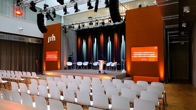 Konferenzraum mit einer Bühne, ausgestattet mit einem Rednerpult und mehreren Stühlen. Im Hintergrund sind orangefarbene Wände mit Beleuchtung und Bildschirmen, die Informationen zur Veranstaltung anzeigen.