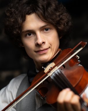 A young man with curly hair is playing the violin. He is wearing a white shirt and a gray vest while looking at the camera. The background is blurred and features dark stones.