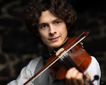 Ein junger Mann mit lockigem Haar spielt Geige. Er trägt ein weißes Hemd und eine graue Weste, während er in die Kamera schaut. Der Hintergrund ist unscharf und zeigt dunkle Steine.