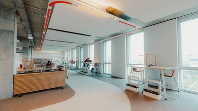 A modern office with large windows, bright workspaces, and an open layout. There are standing desks and seating areas that promote a creative work environment.