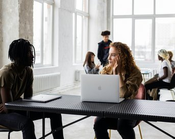 Two people are sitting at a table, engaged in conversation. One person has curly hair and looks thoughtful, while the other, with dreadlocks, listens attentively. In the background, more people can be seen in a modern space.