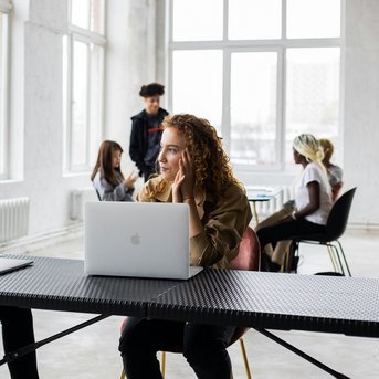 Zwei Personen sitzen an einem Tisch, während sie sich unterhalten. Eine Person hat lockige Haare und schaut nachdenklich, während die andere Person mit Dreadlocks aufmerksam zuhört. Im Hintergrund sind weitere Personen in einem modernen Raum zu sehen.
