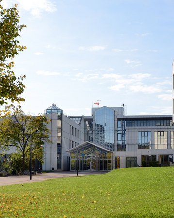 Moderne Gebäude mit Glasfassade und Grünflächen, umgeben von Bäumen und Sträuchern. Ein klarer Himmel ist im Hintergrund sichtbar.
