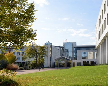 Modern buildings with glass facades and green spaces, surrounded by trees and shrubs. A clear sky is visible in the background.