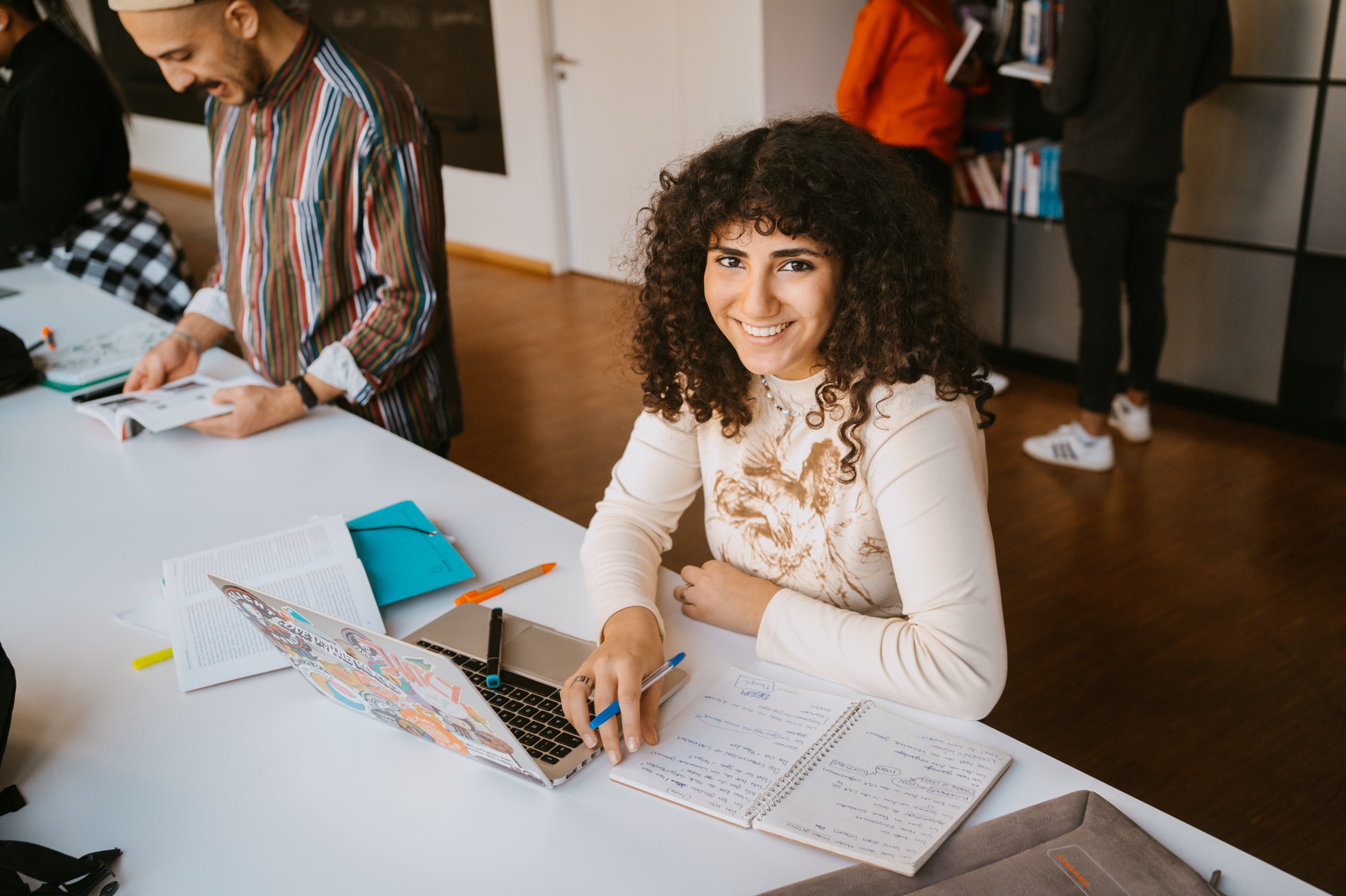 A smiling woman with curly hair sits at a table, with a laptop and notebook in front of her. In the background, there are other people working at another table. The environment feels modern and inviting.