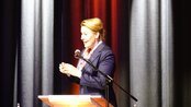 Eine Frau in einem blauen Blazer spricht am Mikrofon, während sie gestikuliert. Im Hintergrund sind dunkle Vorhänge zu sehen.