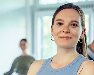 Eine junge Frau mit langen Haaren und Ohrringen lächelt in die Kamera, während zwei Personen im Hintergrund stehen.