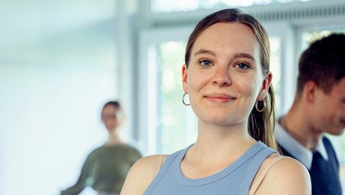 Eine junge Frau mit langen Haaren und Ohrringen lächelt in die Kamera, während zwei Personen im Hintergrund stehen.