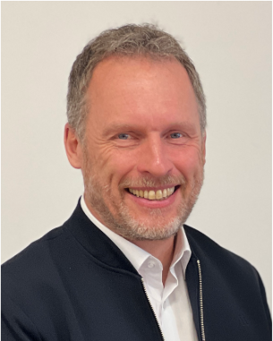 A man with short, gray hair and a beard smiles warmly at the camera, wearing a black jacket over a white shirt.
