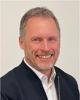 Ein Mann mit kurzem, grauem Haar und Bart lächelt freundlich in die Kamera, trägt eine schwarze Jacke über einem weißen Hemd.