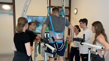 A group of six people is observing and supporting a man who is training to walk with a special device.