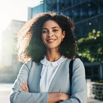 A woman with curly hair stands smiling outdoors, dressed in a gray blazer, with her arms crossed in front of modern buildings.