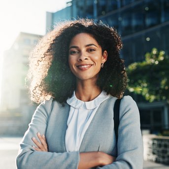 Eine Frau mit lockigem Haar steht lächelnd im Freien, gekleidet in einem grauen Blazer, mit verschränkten Armen vor modernen Gebäuden.