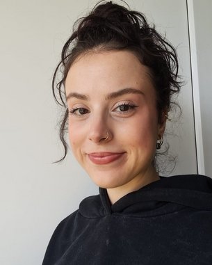 Junge Frau mit lockigem, dunklem Haar und einem Nasenring, trägt einen schwarzen Hoodie und lächelt in die Kamera.