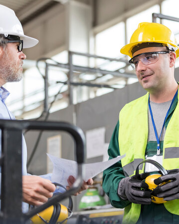 Zwei Männer in Sicherheitskleidung und Helmen diskutieren in einer Industrieumgebung. Der Mann mit dem gelben Helm trägt Gehörschutz.