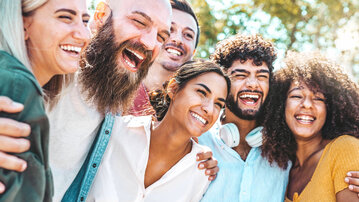 Six people are smiling and laughing together in a sunny setting, embracing each other and sharing joy.