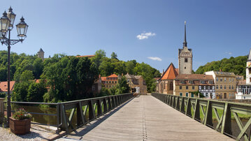 Eine Holzbrücke führt über einen Fluss, umgeben von historischen Gebäuden und Bäumen. Im Hintergrund ist ein hoher Turm und ein klarer blauer Himmel zu sehen.