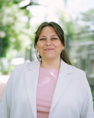 Eine lächelnde Frau mit langen, braunen Haaren trägt einen weißen Blazer und ein rosa Oberteil, steht auf einem Weg umgeben von Pflanzen.