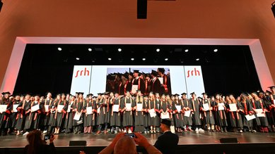 Graduates in academic attire stand on stage, holding their diplomas and posing for photos during a graduation ceremony.
