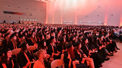 A large group of graduates in black gowns and caps is seated in a festive hall, ready for the graduation ceremony.