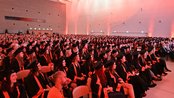 A large group of graduates in black gowns and caps is seated in a festive hall, ready for the graduation ceremony.