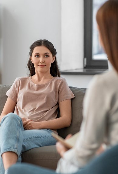 Zwei Frauen sitzen in einem hellen Raum. Eine Frau mit langen, dunklen Haaren trägt ein einfaches T-Shirt und Jeans, während sie der anderen Frau, die ihr gegenüber sitzt, aufmerksam zuhört.