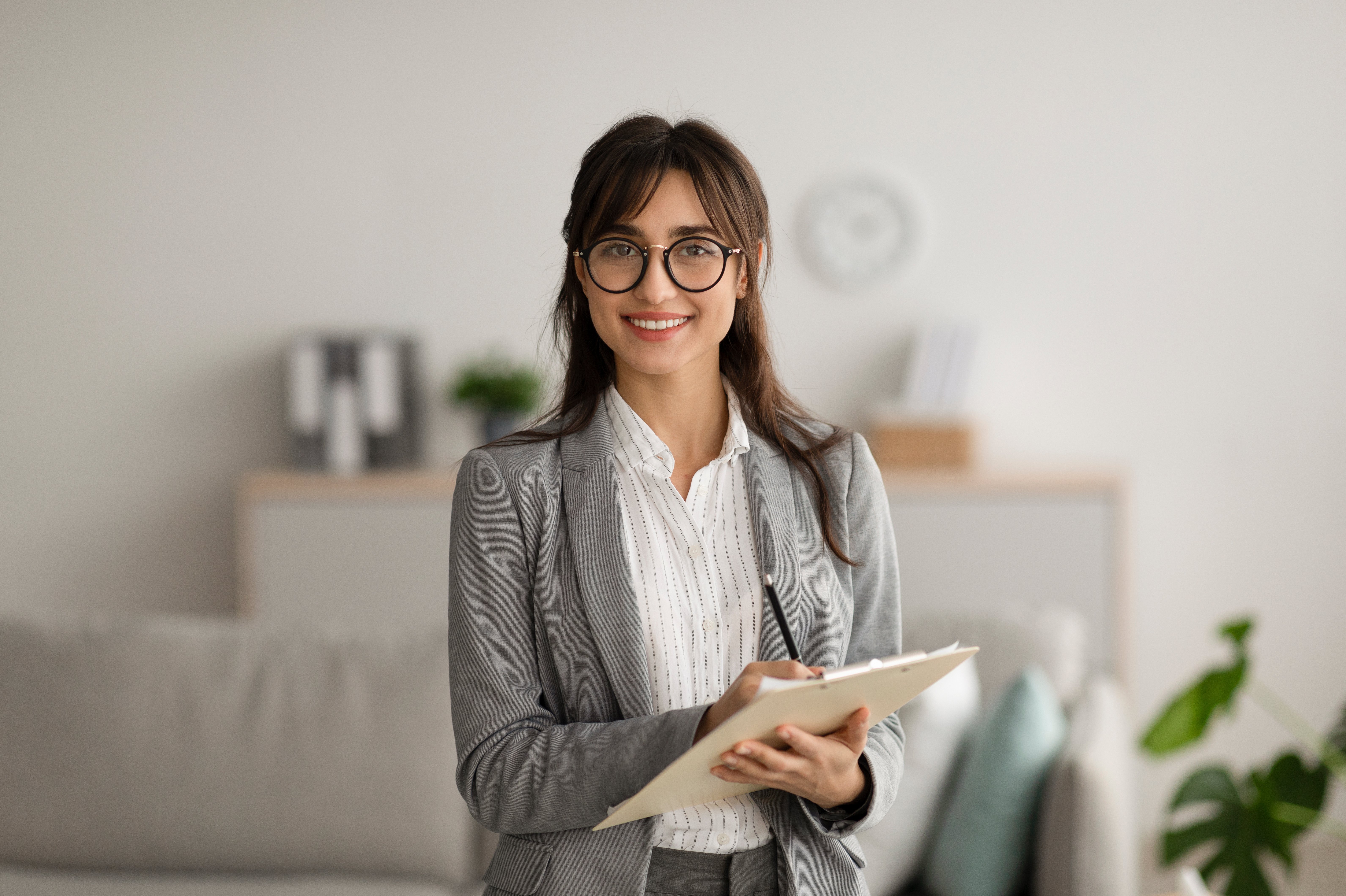 Eine Frau mit Brille trägt einen grauen Anzug und hält ein Notizbuch in der Hand. Sie steht in einem modernen Raum mit neutralen Farben und lächelt freundlich. Pflanzen und Möbel sind im Hintergrund sichtbar.
