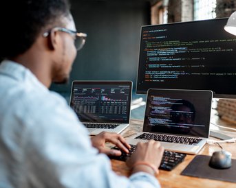 Ein Mann sitzt an einem Schreibtisch und arbeitet an zwei Laptops sowie einem großen Monitor. Auf den Bildschirmen sind Programmiercodes und Datenanalysen sichtbar. Der Raum hat große Fenster und eine moderne Einrichtung.