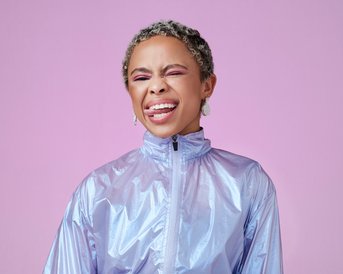 A person with short, curly hair is wearing a shiny silver jacket and smiling while sticking out their tongue. The background is pink.