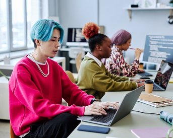 A young man with blue hair is working on a laptop, while two women in a modern office in the background are focused on their tasks.