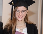 Eine graduierte Frau mit Brille und Doktorhut hält ein Diplom in der Hand, während sie neben einem Mann steht.