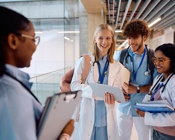 Vier Medizinstudenten in weißen Kitteln unterhalten sich in einem modernen Flur, während sie Tablets und Unterlagen halten.